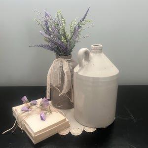 May include: A white ceramic pitcher with a handle sits on a lace doily next to a brown ceramic vase with a burlap ribbon. The vase holds a bouquet of purple and green flowers.
