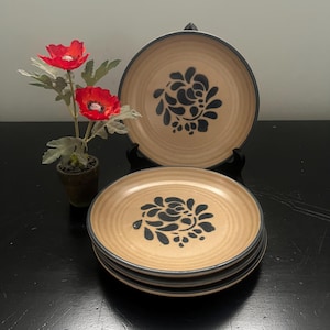 May include: Set of tan-colored ceramic plates with a dark blue floral design in the center and a dark blue rim. The plates are stacked and displayed with a decorative flower arrangement.