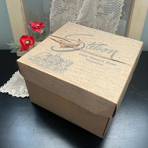 May include: A tan Stetson box with a textured surface, tied with twine. The box features the Stetson logo and address: 1224 Chestnut Street, Philadelphia. The box is on a black surface with red flowers and lace in the background.