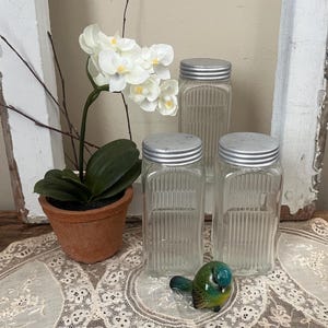 May include: Three clear glass jars with silver lids, arranged on a lace tablecloth. A small ceramic bird and a potted orchid are also present. The jars have vertical ribbed detailing.