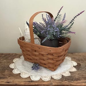 May include: A brown wicker basket with a handle, filled with lavender flowers and a white fabric item. The basket is sitting on a white doily on a wooden surface.