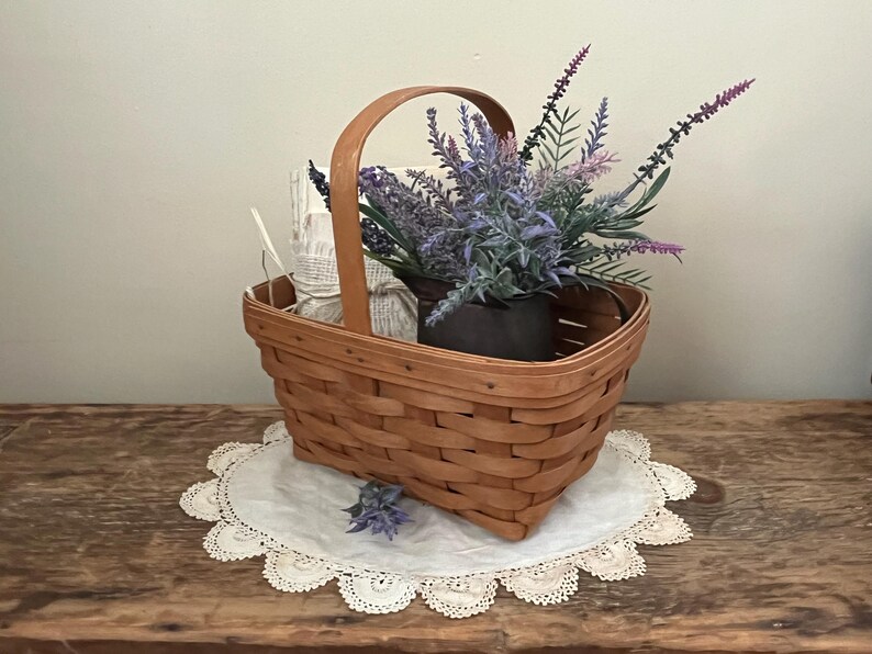 May include: A brown wicker basket with a handle, filled with lavender flowers and a white fabric item. The basket is sitting on a white doily on a wooden surface.