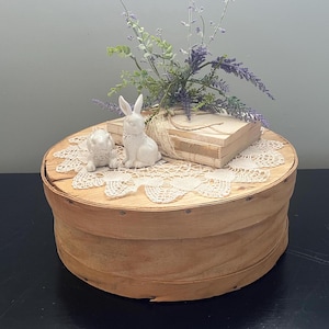 May include: A wooden round box with a white lace doily and two white porcelain bunny figurines. The box is decorated with a bouquet of purple flowers.