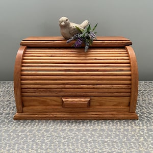 May include: A wooden bread box with a roll-top lid and a small drawer. The box is made of light brown wood and has a decorative bird figurine and purple flowers on top. The bread box is designed to store bread and baked goods.
