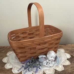 May include: A woven wood basket with a wooden handle. The basket is sitting on a white crocheted doily with a porcelain angel figurine and purple flowers.