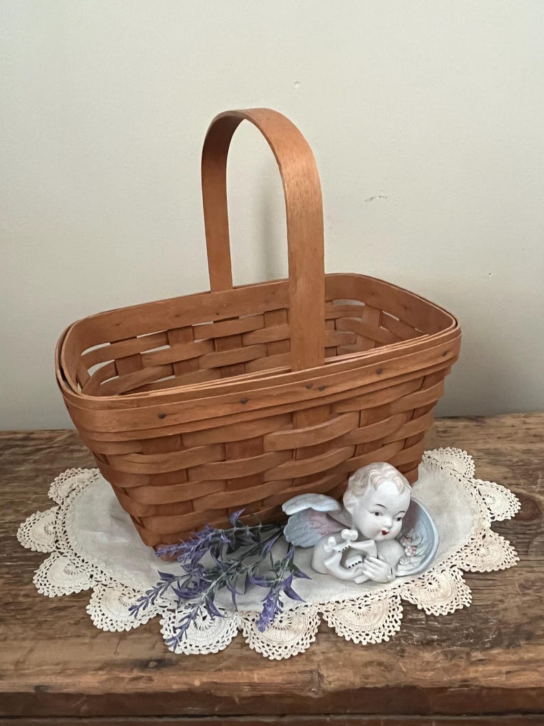 May include: A woven wood basket with a wooden handle. The basket is sitting on a white crocheted doily with a porcelain angel figurine and purple flowers.