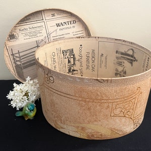 May include: A vintage hat box with a brown and white patterned paper exterior. The box is open and the inside is lined with a newspaper print. The newspaper print includes advertisements for furniture and paintings. The box is sitting on a black surface.