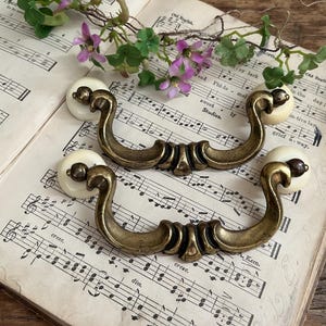 May include: Two antique brass drawer pulls with cream-colored ceramic knobs. The pulls are curved with ornate detailing and are set on an open book of sheet music, with sprigs of greenery and purple flowers.