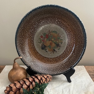May include: A brown ceramic plate with a speckled glaze and a painted design of fruit in the center. The plate is on a black stand and a white tablecloth.