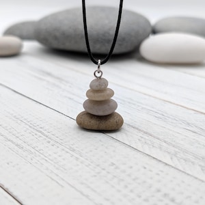 May include: A pendant necklace with a stack of five smooth, natural stones in shades of brown and beige. The stones are suspended from a black cord with a silver-coloured metal loop.