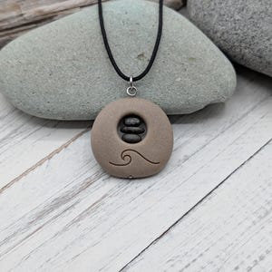 Op de afbeelding: Een beige, ronde stenen hanger met een gegraveerd golfpatroon en een centrale uitsparing met drie kleine, gestapelde donkergrijze kiezelstenen. De hanger hangt aan een zwart koord en vormt zo een ketting met een natuurlijke uitstraling.