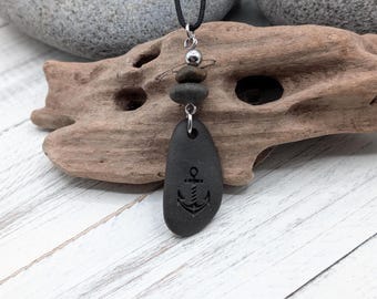 Puede incluir: Un collar con cordón negro y un colgante formado por tres piedras grises apiladas. La piedra inferior, la más grande, tiene grabado un símbolo de ancla negra. Una pequeña cuenta de metal plateado se sitúa entre las dos piedras superiores.