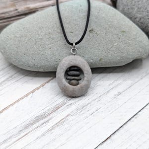 Op de afbeelding: Een ketting met een hanger bestaande uit een ronde grijze steen met een gat in het midden, waarin een kleine stapel van drie donkere, gladde kiezels zit. De hanger hangt aan een dun zwart koord met een zilverkleurige ring.