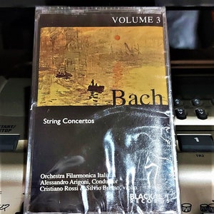 May include: A cassette tape with a black and gold label featuring a painting of a cityscape at sunset. The text "Bach" and "String Concertos" are printed on the label. The cassette tape is labeled "Volume 3" and is part of a series of recordings by the Orchestra Filarmonica Italiana.