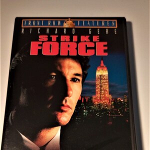 Può includere: Custodia DVD del film "Strike Force" con Richard Gere. La copertina presenta il ritratto di Gere, il titolo in rosso e un paesaggio urbano notturno con un grattacielo illuminato. La custodia del DVD è nera.