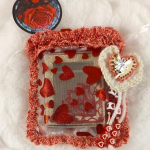 May include: A small red and white crocheted pouch with a clear plastic window. Inside the pouch is a small book with a red glitter heart design. The pouch also has a white beaded charm with a heart and a small silver fairy.