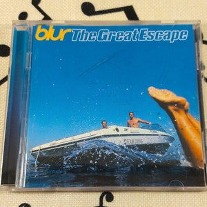 以下が含まれることがあります： 青い背景に「blur The Great Escape」という黄色の文字が書かれたCDケース。画像には、水上のボートに乗った3人の人物が写っており、1人の人物の足が空中に突き出ている