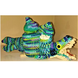 May include: A knitted green, blue and purple creature with a large mouth and white teeth. The creature is holding a container of cotton buds.