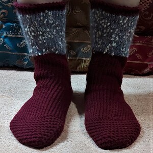 May include: A pair of handmade knitted socks. The socks are primarily a deep burgundy color, with a textured knit pattern. The cuffs are a speckled gray and white, with a burgundy trim. The socks are shown on a person's feet.