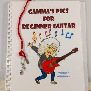 Op de afbeelding: Een wit spiraalgebonden boek met een rood gehaakt bladwijzer. De omslag toont een cartoonillustratie van een vrouw die een rode elektrische gitaar bespeelt met muzieknoten rond haar hoofd. De titel van het boek is "GAMMA'S PICS FOR BEGINNER GUITAR".