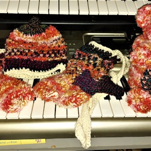 May include: A set of crocheted items including a hat, mittens, and a teddy bear. The hat and mittens are multi-coloured with shades of pink, red, and white. The teddy bear is pink and white with black eyes and a black nose.