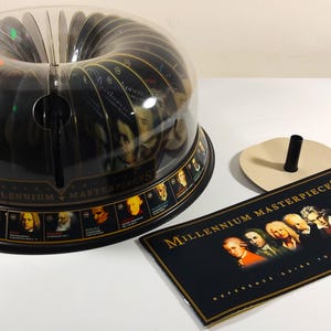 May include: A black and clear plastic CD holder with a clear dome, containing multiple CDs. The base has portraits of historical figures. A reference guide and a stand are next to the holder. The text "Millennium Masterpieces" is visible.