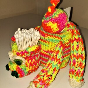 May include: A crocheted cotton bud holder shaped like a person wearing a bright pink, yellow and green sweater. The holder has a small pocket on the front for the buds.