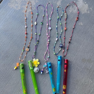 May include: Five reusable plastic tubes in green, blue, and red, each attached to a colorful beaded lanyard. The lanyards feature various charms including a yellow smiley face, a disco ball, and evil eye beads. The tubes are labeled Reutilizable.