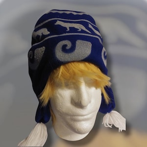 Puede incluir: Sombrero de forro polar azul con un patrón blanco de animales y espirales. El sombrero tiene pompones blancos en los extremos de las orejeras.