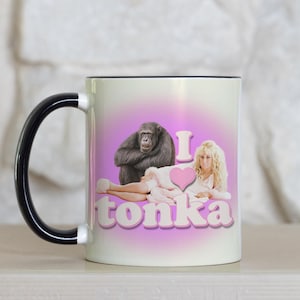 May include: White ceramic coffee mug with a black handle. The mug features a pink background with the text "I love Tonka" and an image of a chimpanzee and a woman.