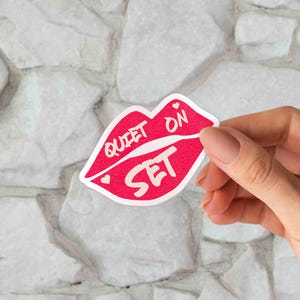 May include: A red sticker in the shape of lips with the words "QUIET ON SET" in white. The sticker has a white outline and two small hearts. The sticker is held against a white stone background.