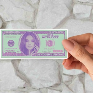 May include: A novelty $100 bill featuring a portrait of Selena Gomez. The bill is printed on a light green background with purple text and a purple border. The text reads "The United States of Selena" and "One Hundred Dollars".