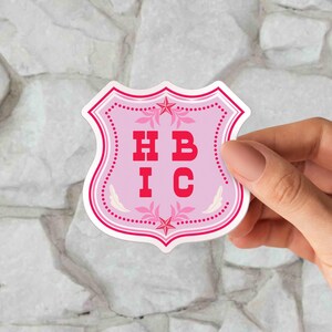 May include: A pink sticker with a shield shape, featuring the letters "HBIC" in red. The sticker has a decorative border with stars and floral accents. The sticker is held by a hand against a stone background.