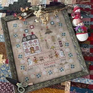 May include: Framed cross-stitch artwork with a winter theme. The design features a house, snowflakes, and the word "WINTER". The frame is ornate with a dark, antique finish. A pair of scissors and two snowman figurines are also present.
