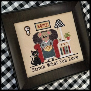 May include: Cross-stitch embroidery of a mouse sitting in a red armchair with a black cat beside it. The mouse is wearing a grey dress and holding a yellow ball. The text "Stitch What You Love" is below the mouse and cat. The embroidery is framed in a dark brown frame.