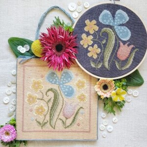 May include: Two embroidery patterns featuring floral designs with blue, pink, yellow, and green flowers. The patterns are stitched on cream-colored fabric and framed in wooden hoops.
