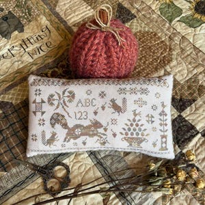 May include: A rectangular cream-colored fabric pincushion featuring cross-stitch embroidery of squirrels, acorns, the letters ABC, and the numbers 123, accompanied by a round, ribbed, dark orange yarn pumpkin with a cinnamon stick stem.