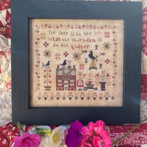 May include: A framed cross-stitch sampler with a beige background and colorful embroidery. The sampler features a small house, birds, and a quote: "The skep is to the bee what the churn dash is to the quilter."