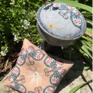 May include: Two small needlepoint pincushions with butterfly designs. One is light blue with a single butterfly and the other is peach with a repeating butterfly pattern. Both are on a wooden stand.