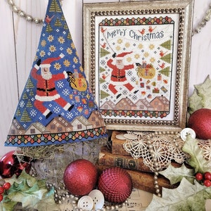 May include: A framed cross-stitch design featuring a Santa Claus figure with a bag of gifts, surrounded by a snowy landscape and the text "Merry Christmas". The design is displayed on a table with a blue and white Christmas tree ornament, red ornaments, and a string of pearls.