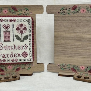 May include: Wooden cross-stitch display stand with a stitched design that reads "A Stitcher's Garden." The stand has a light brown finish with floral accents in pink, green, and burgundy. Includes a small wooden thread holder.