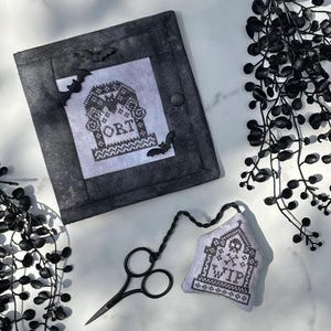 May include: A black and white cross-stitch project with a tombstone design, the word "ORT" is visible. A small, gray, stitched bag with the letters "WIP" is attached to a pair of black scissors. Decorative black bats and foliage complete the Halloween theme.