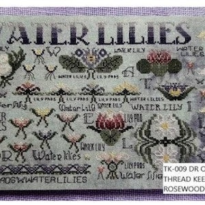May include: A cross-stitch design featuring the words "WATER LILIES" and various embroidered water lily motifs. The design includes text such as "WATER LILY" and "LILY PADS". The piece is mounted on a light gray, rectangular frame with circular cutouts.