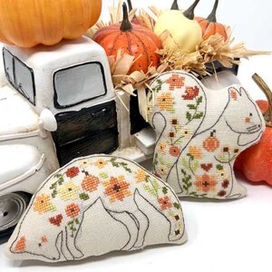 May include: Two white fabric squirrels with embroidered floral patterns in shades of orange, yellow, and green. The squirrels are sitting on a white toy truck bed filled with hay and pumpkins.