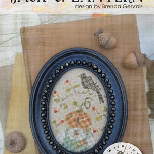 May include: An oval black frame with a beaded edge containing a cross-stitch embroidery of a pumpkin character with a green stem, a bird, and the number 6, surrounded by small floral motifs on a light fabric background.
