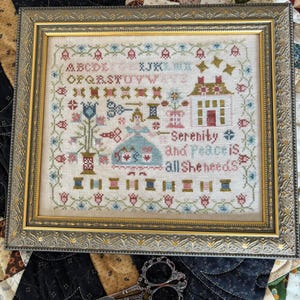 May include: A framed cross-stitch sampler with a gold-colored ornate frame. The sampler features a house, flowers, and the text "Serenity is all She needs." Decorative scissors are in the foreground.