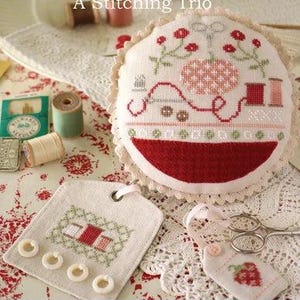 May include: A round pincushion with cream lace trim, a rectangular tag with four white buttons, and a small strawberry-themed tag. The items feature cross-stitch designs of sewing supplies, including spools, scissors, and a thimble.