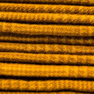 May include: Close-up of folded, mustard-yellow fabric. The fabric has a textured, woven appearance with visible horizontal lines. The color is a warm, earthy tone, suggesting a cozy textile. The image highlights the detailed weave and the fabric's overall texture.
