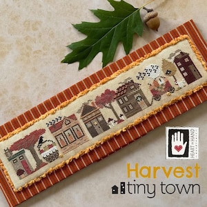 May include: Cross-stitch pattern of a row of houses with autumnal details, including trees, pumpkins, and a witch's hat. The pattern is framed in a brown and orange striped fabric with a fringe border. The text "Harvest tiny town" is below the pattern.