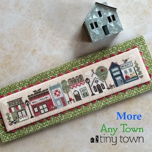 May include: A cross-stitch embroidery of a small town with red, green, and blue buildings. The buildings have names like "Holly Shopping" and "Whimsy". The embroidery is framed with a green fabric with a scalloped edge.
